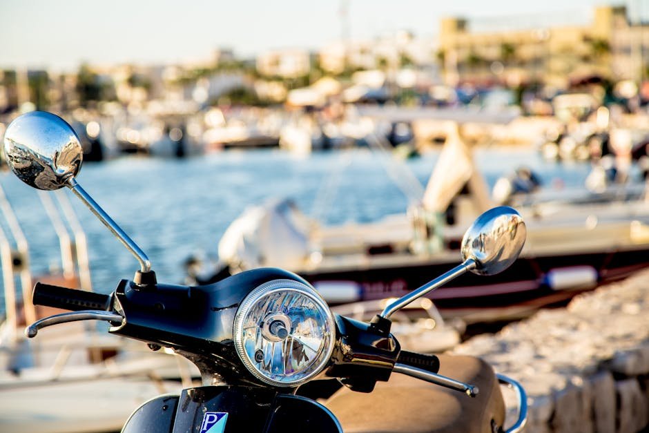 Close-up view of a classic scooter with boats in background at a sunlit seaside marina.